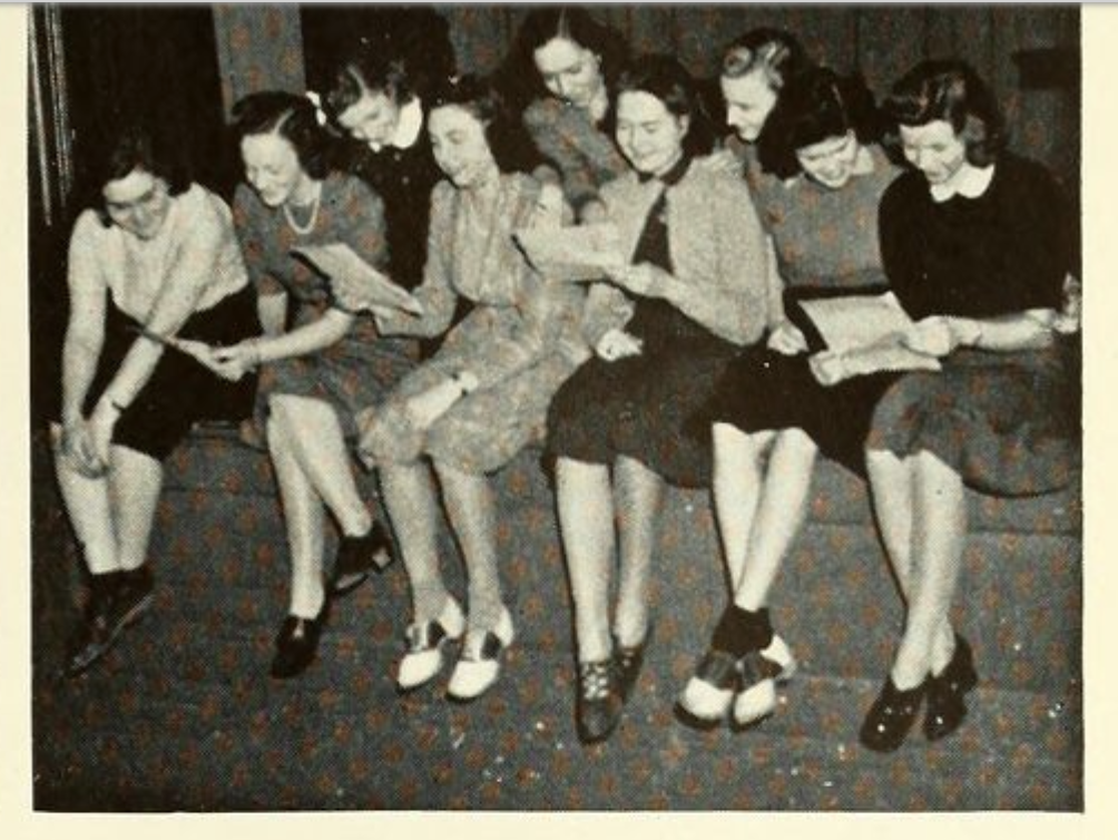 About a dozen young women sit on the edge of a stage rehearsing