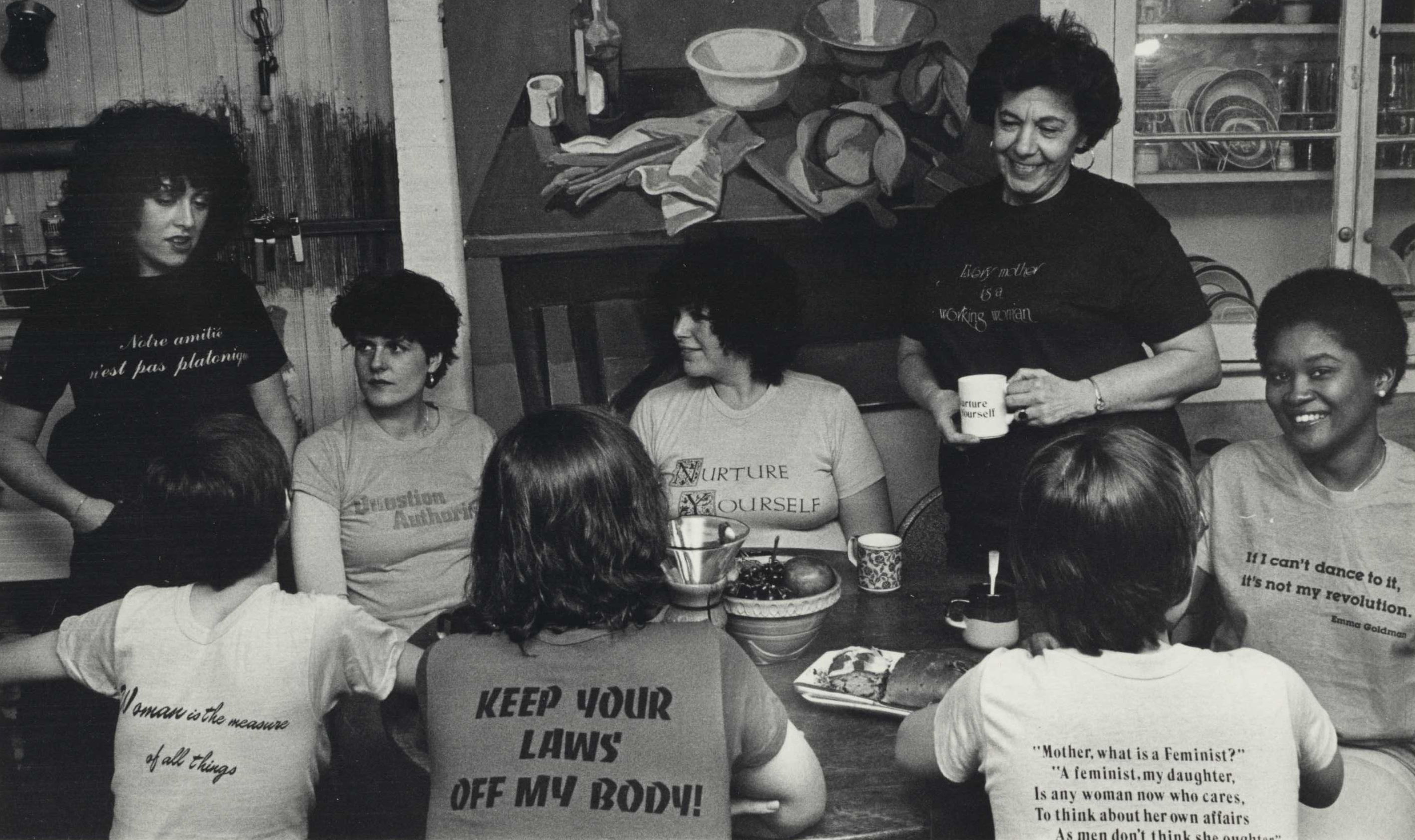 Multiple women wearing graphic tees bearing feminist statements gather around a table