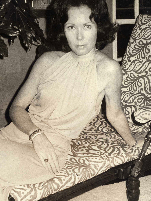 Nancy Friday reclines on a lounge in an evening gown, legs crossed, one hand with a large ring draped seductively across her legs. She boldly stares into the camera.